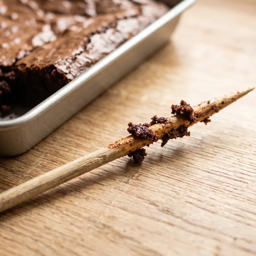 A wooden skewer withdrawn from the centre of a pan of brownies, carrying several moist dark crumbs rather than wet liquid batter.